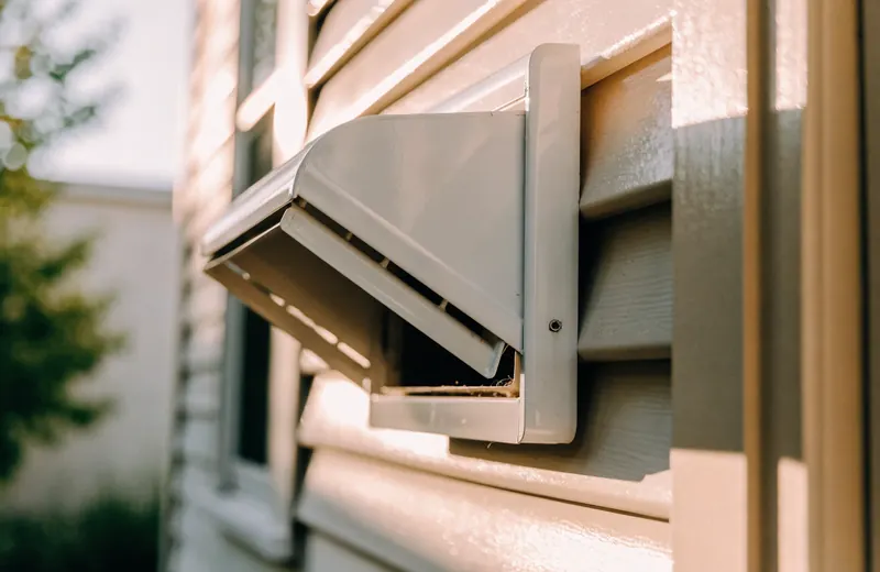 A clothes dryer exhaust vent hood with louvers on the exterior siding of a house
