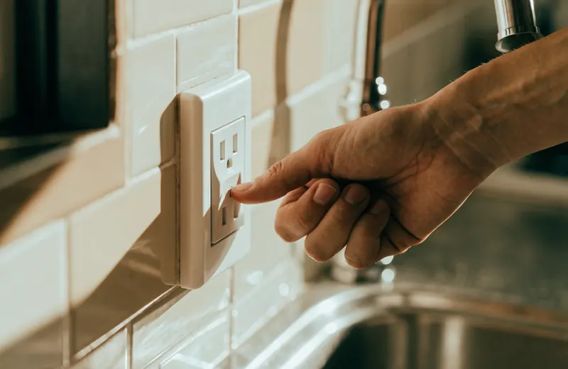 A hand pressing the test button on a GFCI outlet beside a kitchen sink in a bright A hand pressing the test button on a GFCI outlet beside a kitchen sink in a bright, sunlit kitchen